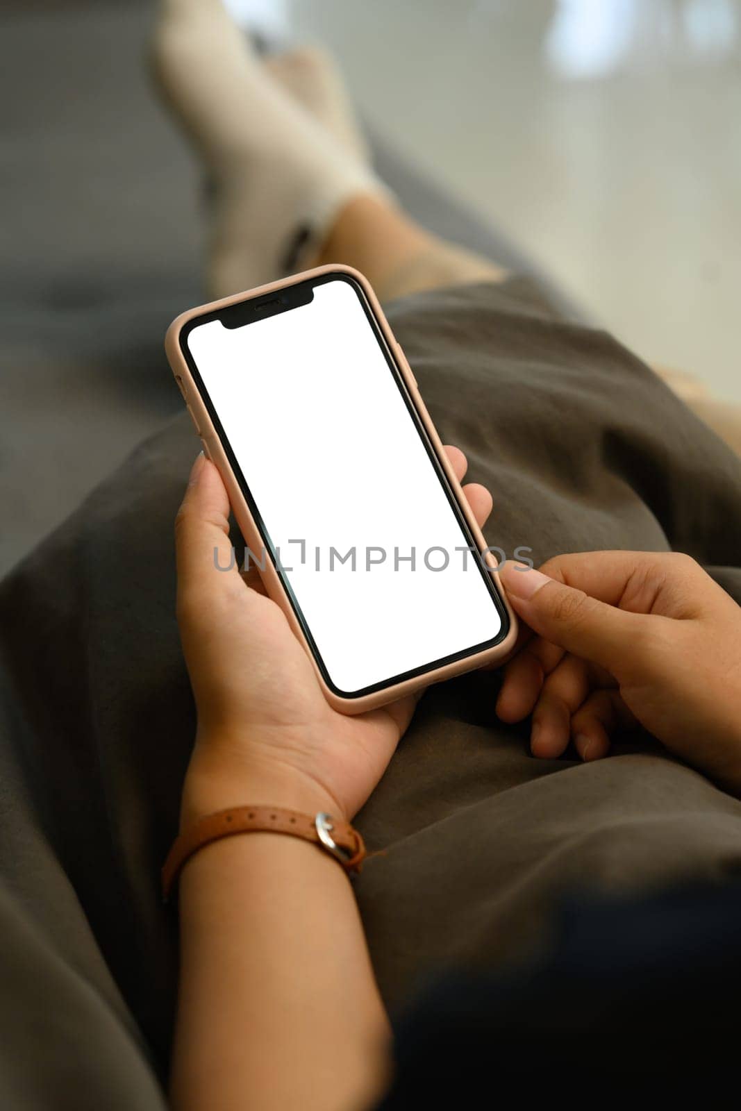 Close up of hands holding a smartphone with white screen while relaxing on sofa.