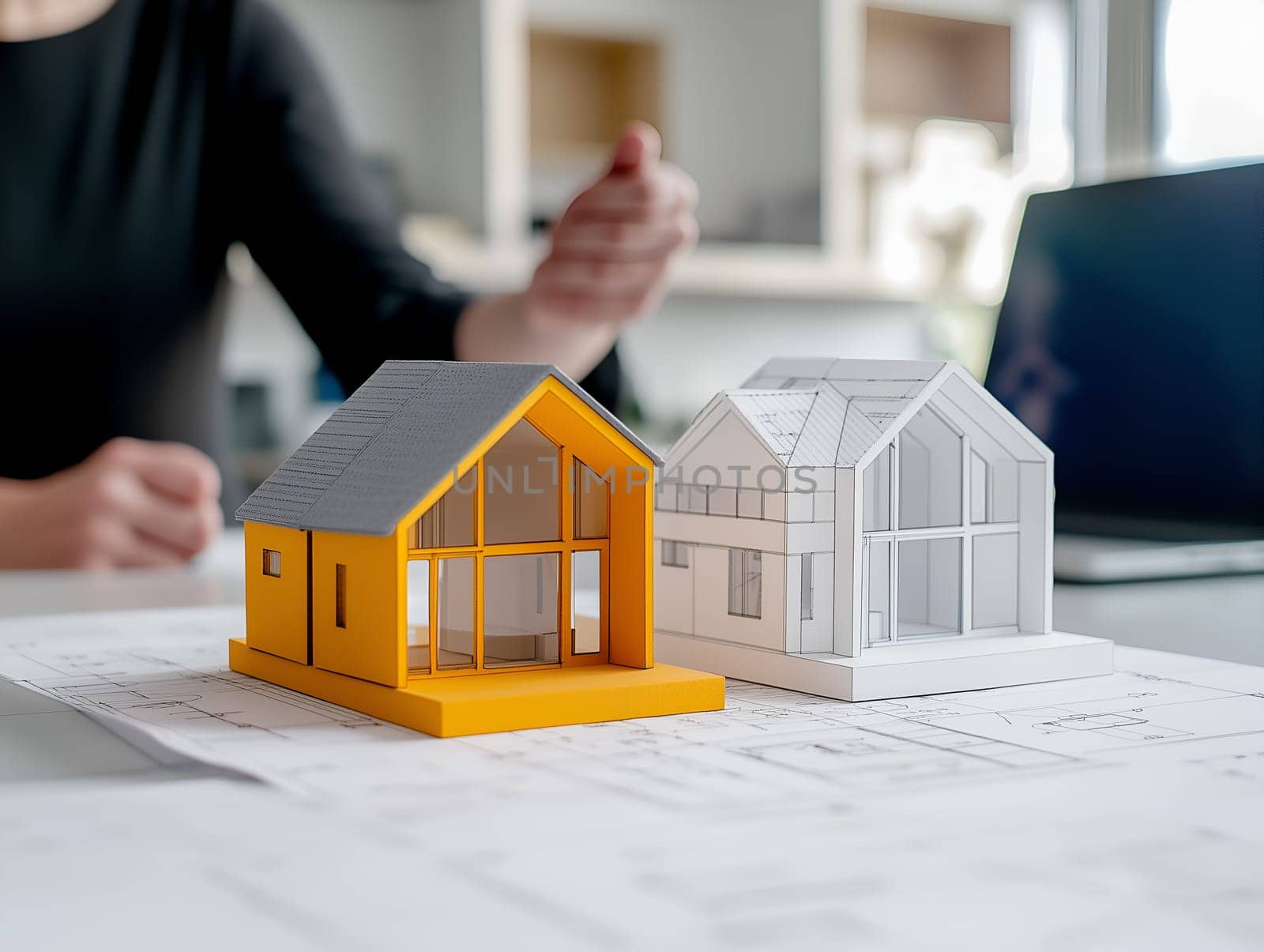Two detailed house models, one in yellow and one in white, placed on blueprints. A person in the background gestures while discussing architectural designs in a modern office.