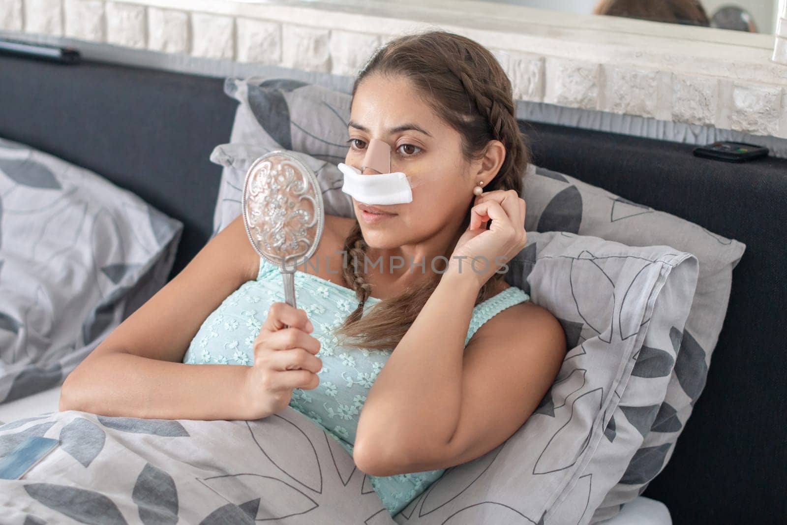 a woman lies in bed in her home clothes with bandages on her face after a nose operation and looks at her reflection in the mirror, adjusts her hair, and worries about the result,