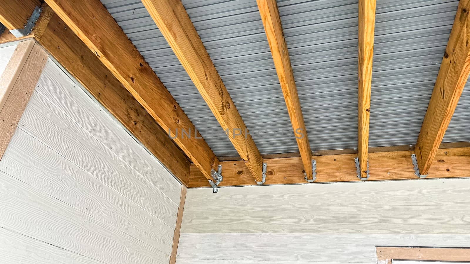 Exposed wood beam ceiling structure supporting a corrugated metal roof in a covered outdoor patio area of a newly built residential home. The design showcases natural wood framing and metal roofing components, providing durability and modern aesthetics.