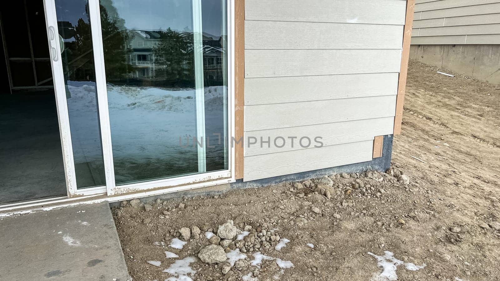 Under-construction backyard patio area featuring a sliding glass door leading to a small concrete step surrounded by dirt and snow. The unfinished landscaping and exposed ground highlight the early stages of residential development.