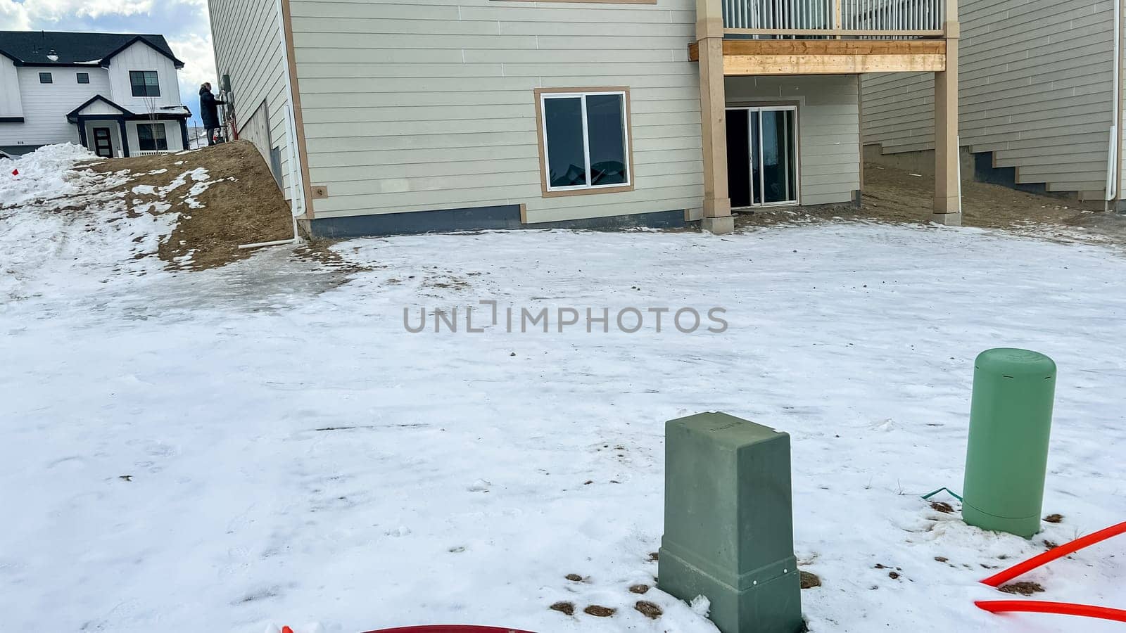 Castle Pines, Colorado USA - January 18, 2023-Rear view of a residential home under construction in a developing neighborhood. The backyard features snow covered ground, utility boxes, and unfinished landscaping. Sliding glass doors lead to the basement, while the upper deck adds to the home modern design.