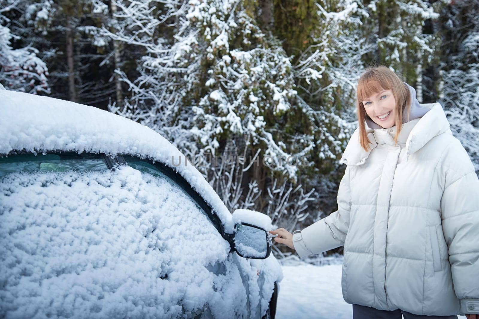 A woman in a warm, puffy jacket stands next to a snow-covered car, enjoying a winter walk in a serene snowy forest landscape.