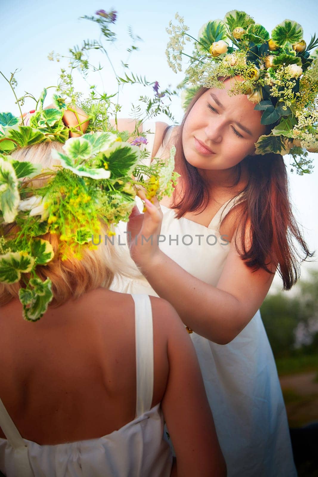 Mother and daughter in white sundresses, wreaths of flowers and bouquet in nature evening at sunset . Family celebrate Slavic Holiday of Ivan Kupala