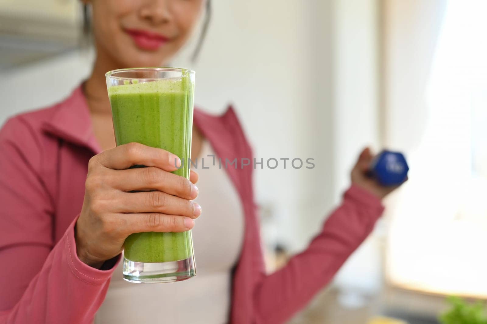 Sporty young woman holding glass of green detox vegetable juice and lifting dumbbell. Dieting and wellbeing concept by prathanchorruangsak