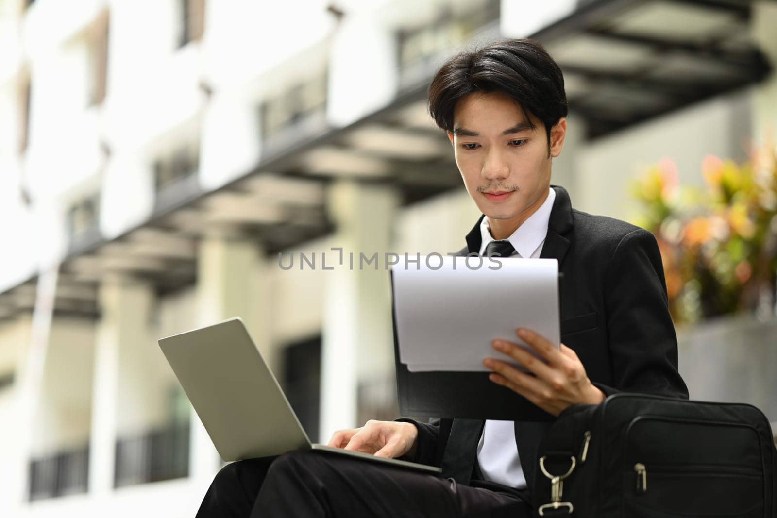 Businessman in formalwear using laptop and reviewing document while sitting in front of business center by prathanchorruangsak
