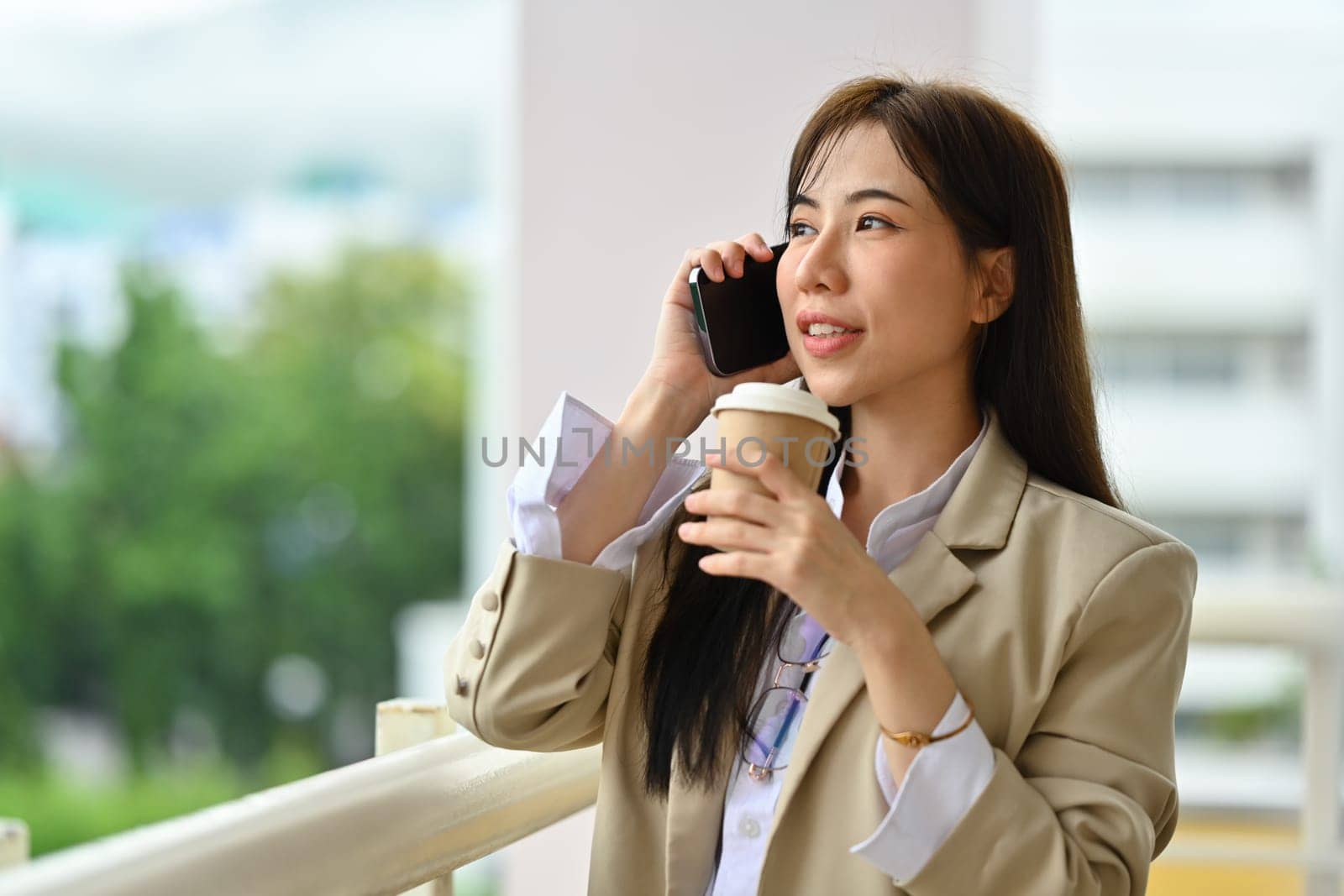 Attractive millennial businesswoman drinking coffee and talk mobile phone at office terrace.