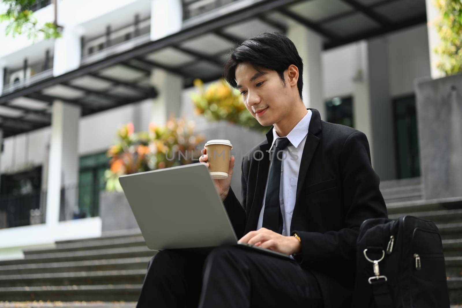 Attractive young businessman sitting on stairs and checking email on laptop before going to the office by prathanchorruangsak