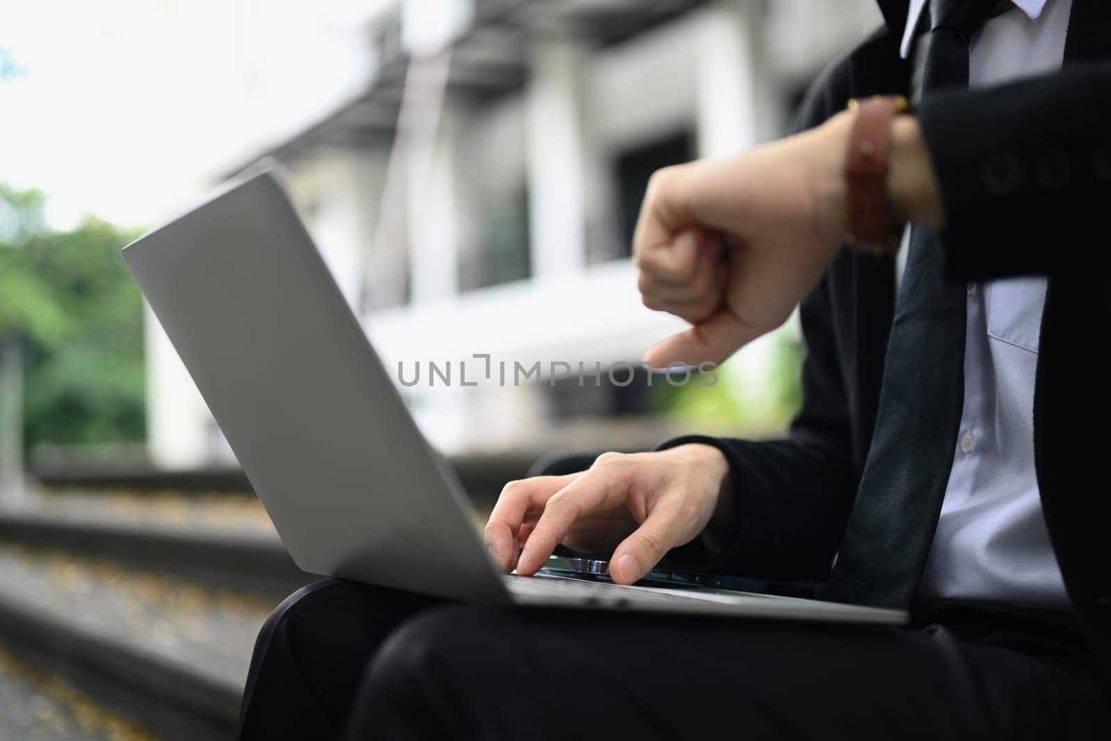 Cropped shot of businessman using laptop and checking time on wristwatch for appointment or transportation by prathanchorruangsak