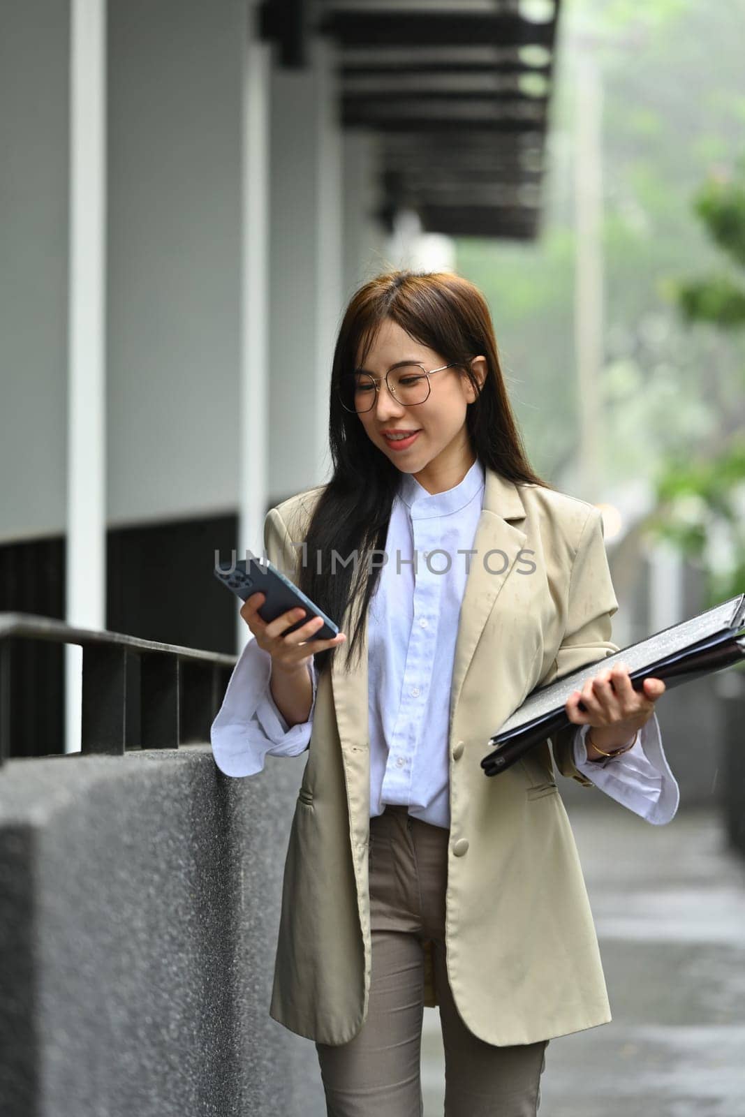 Beautiful female entrepreneur walking near office building chatting online with friends on mobile phone.