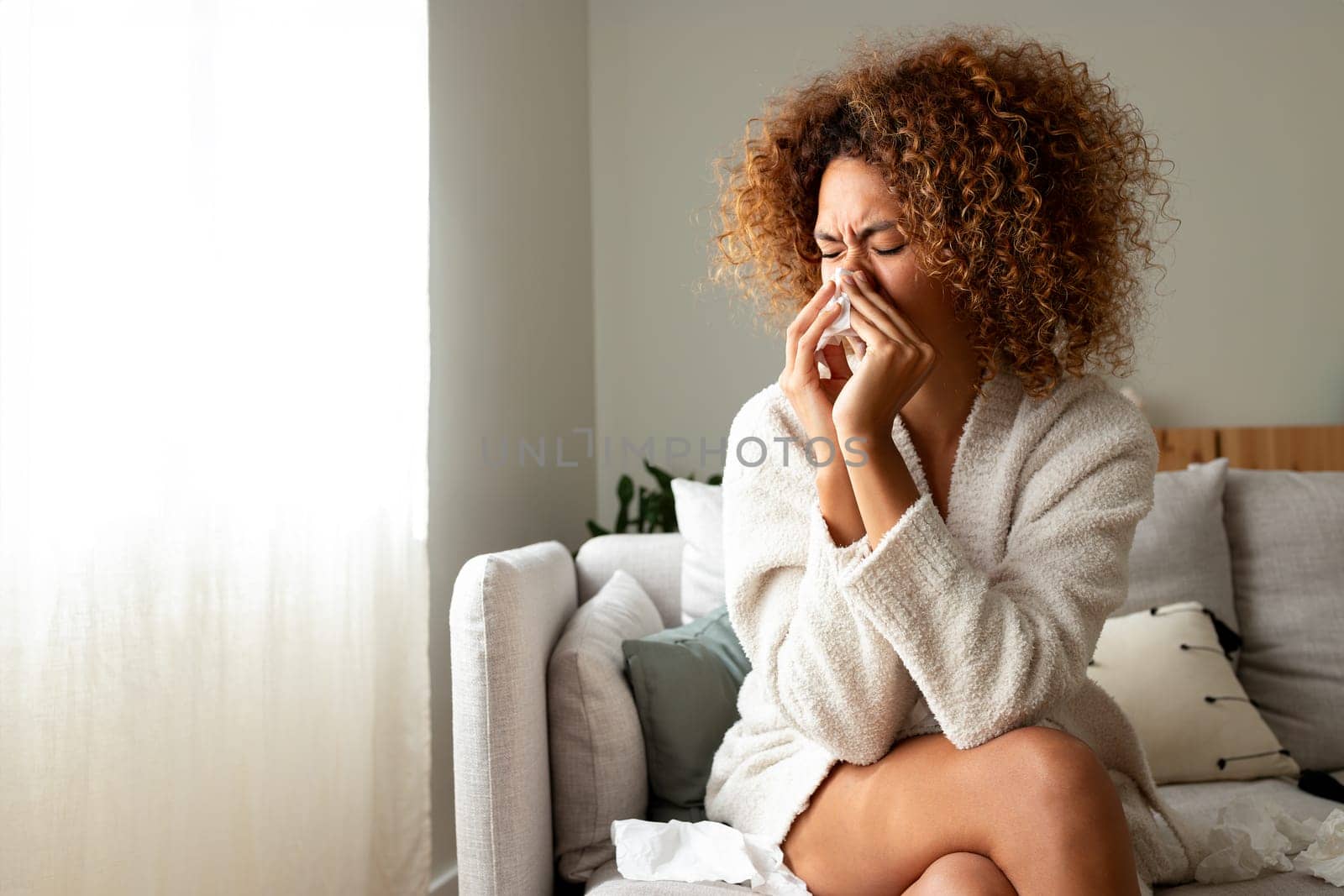 Sick multiracial hispanic woman with the flu blowing nose with tissue paper at home sitting on the sofa. Copy space. by Hoverstock