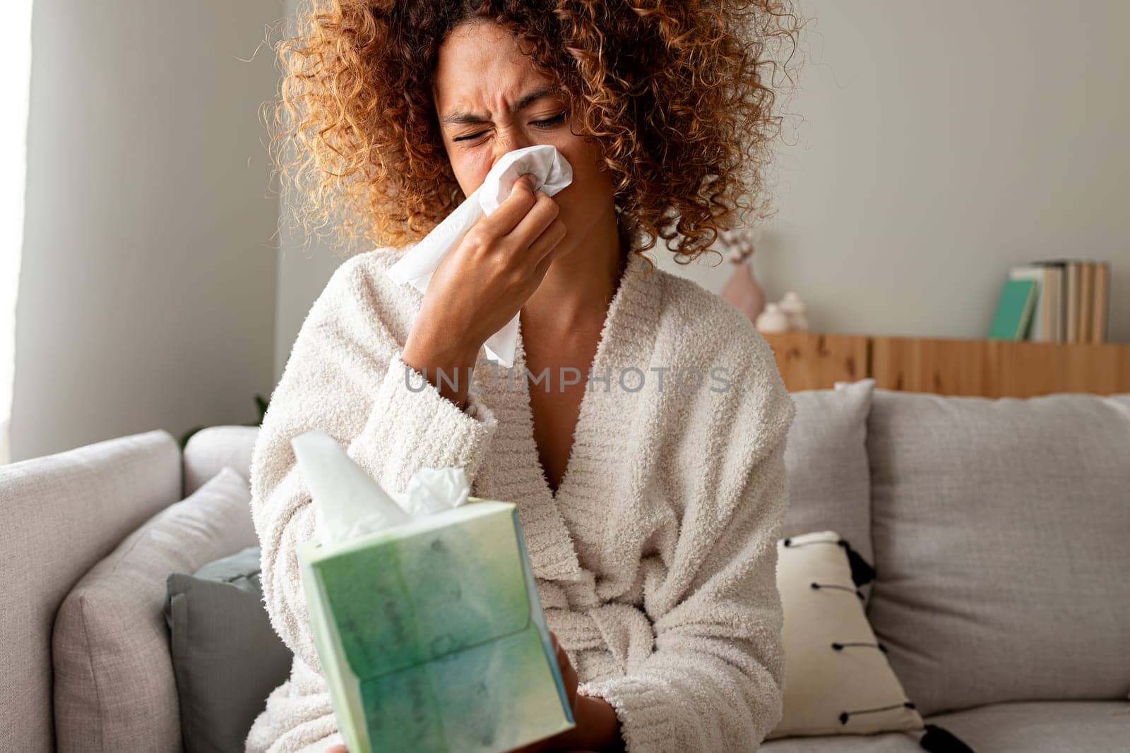 Sick young multiracial woman blowing nose with tissue paper at home sitting on the sofa. Sickness concept.