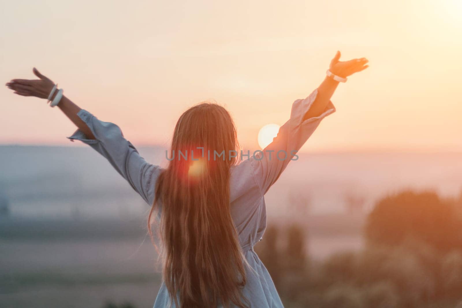 Happy woman standing with her back on the sunset in nature in summer with open hands. Romantic beautiful bride in white boho dress posing with mountains on sunset by panophotograph