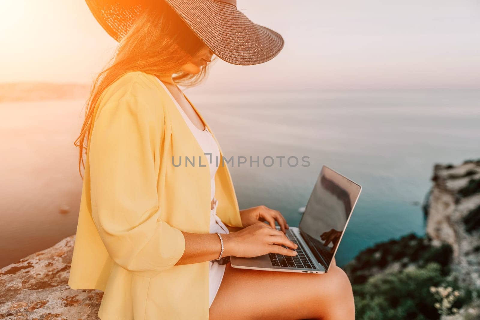 Digital nomad, Business woman working on laptop by the sea. Pretty lady typing on computer by the sea at sunset, makes a business transaction online from a distance. Freelance, remote work on vacation by panophotograph