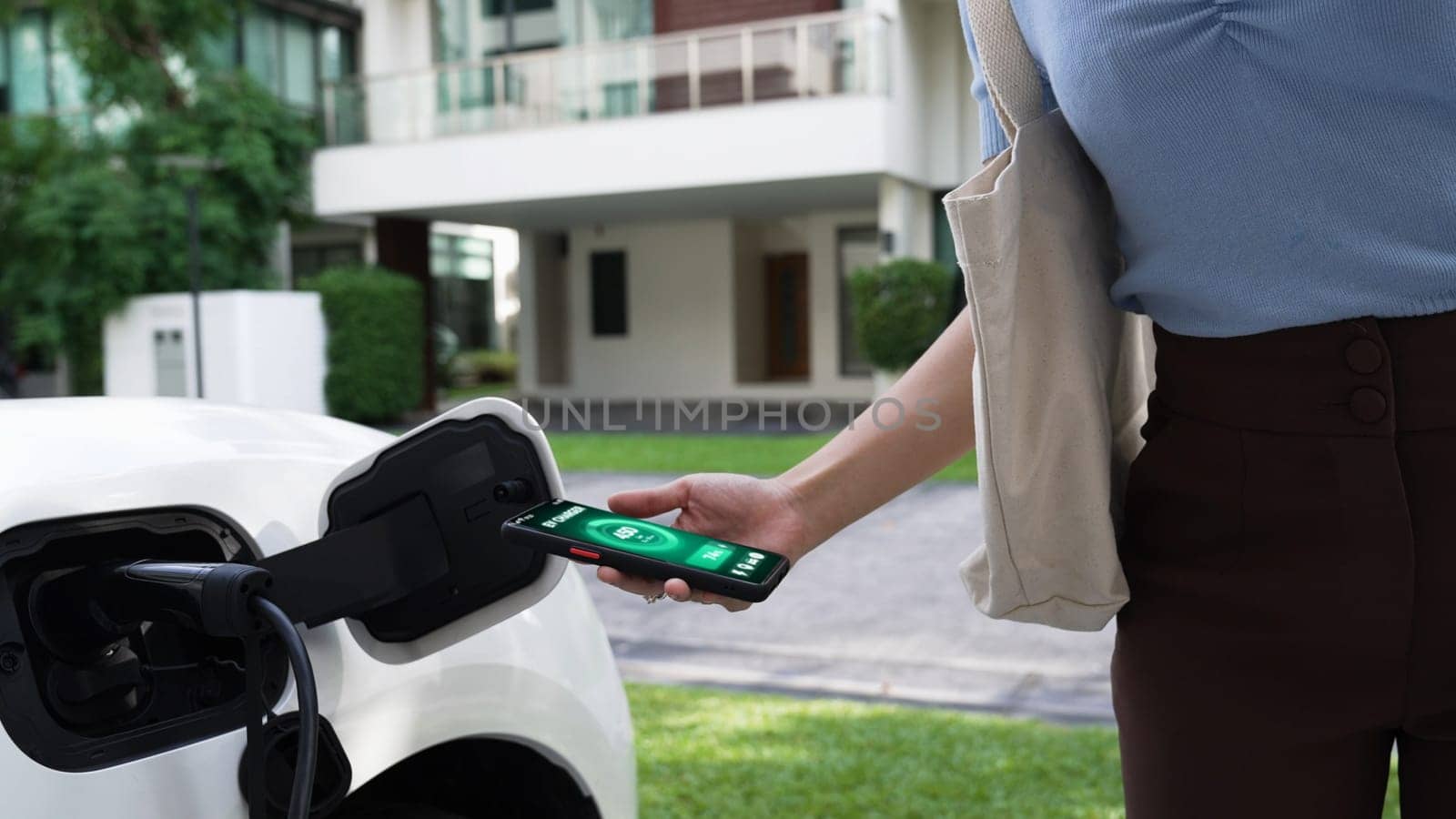 Modern woman recharge her EV car from home charging station by EV charger, checking battery status from EV smartphone app. Future lifestyle of sustainable clean energy utilization. Peruse