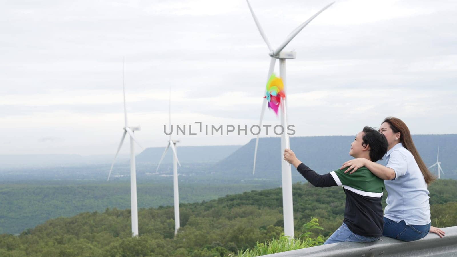 Progressive happy mother and her son at the wind turbine farm. Electric generator from wind by wind turbine generator on the country side with hill and mountain on the horizon.