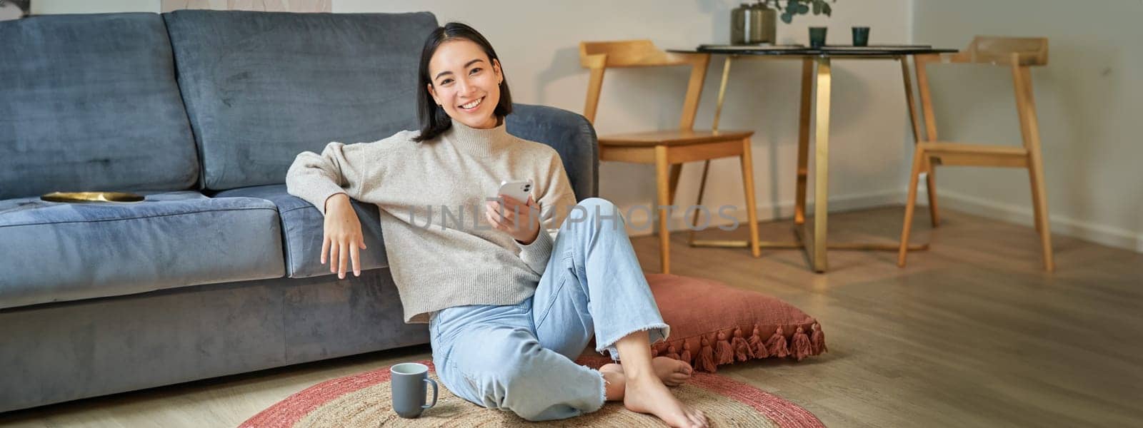 Portrait of stylish korean woman sits on floor with smartphone, using mobile phone, smiling pleased, concept of staying at home and relaxation by Benzoix
