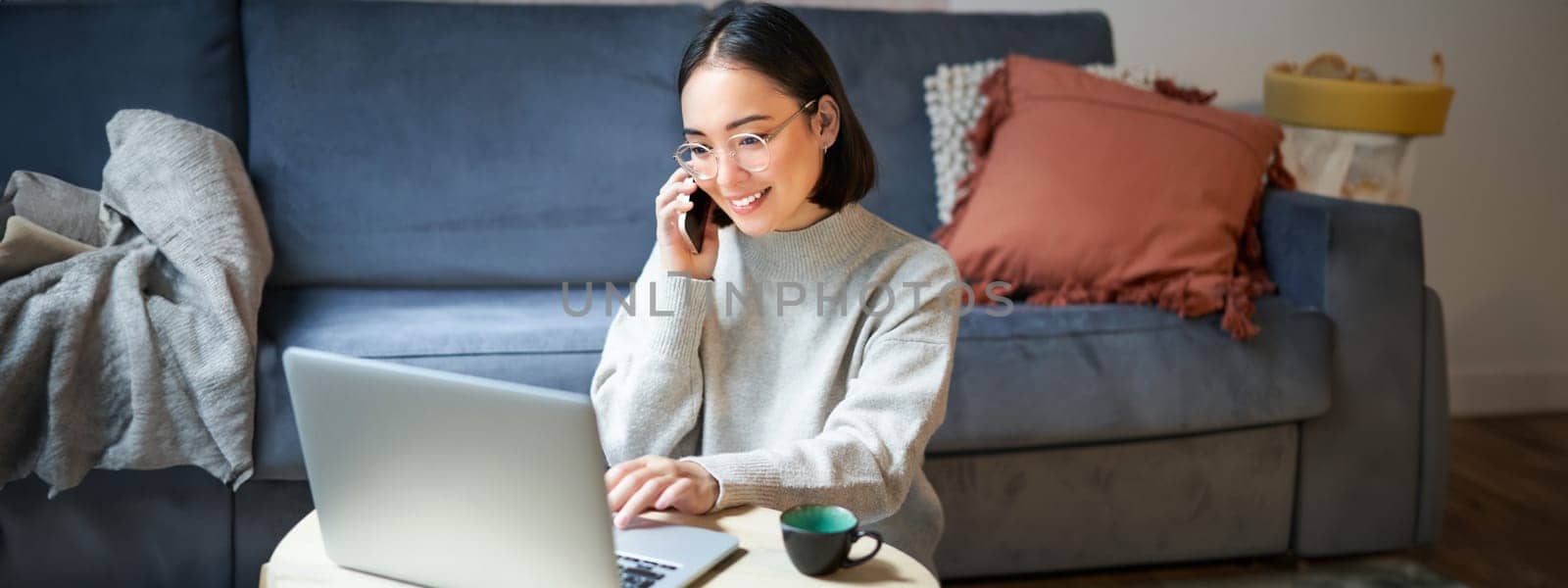 Portrait of smiling asian woman making phone call, working on laptop and having conversation on smartphone.