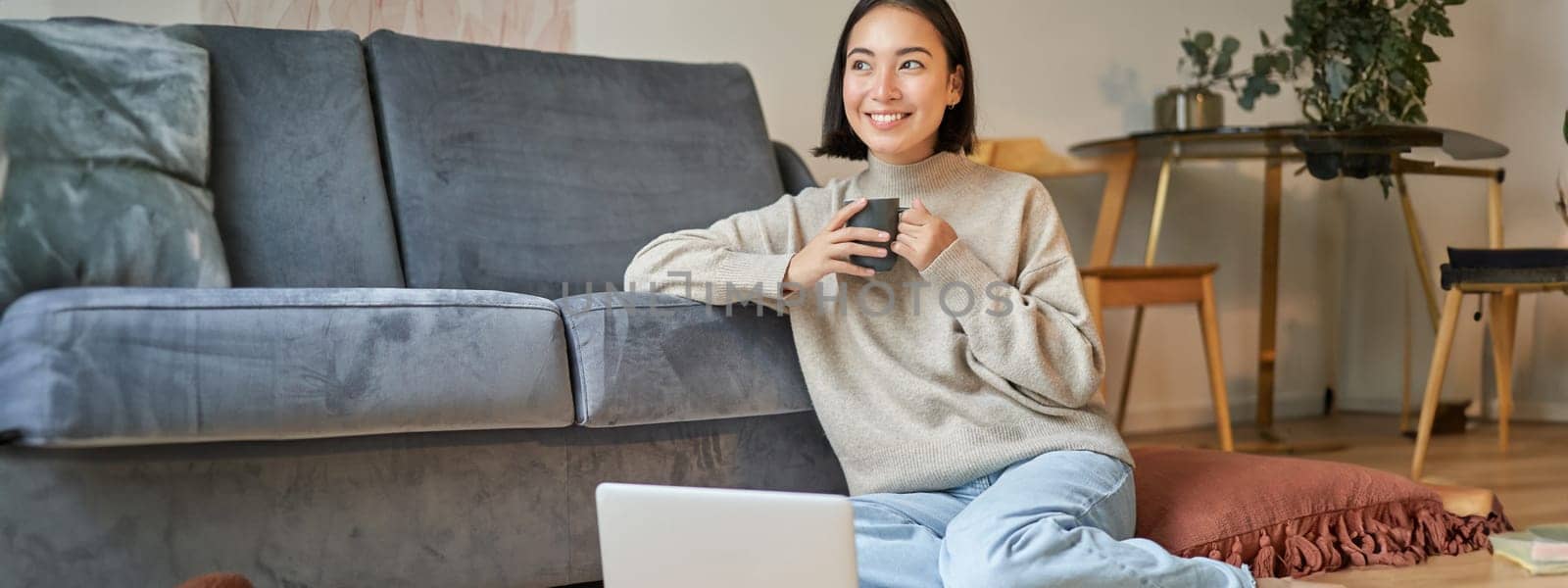 Image of smiling asian woman drinking hot tea, holding cup and sitting near laptop on floor, resting at home by Benzoix