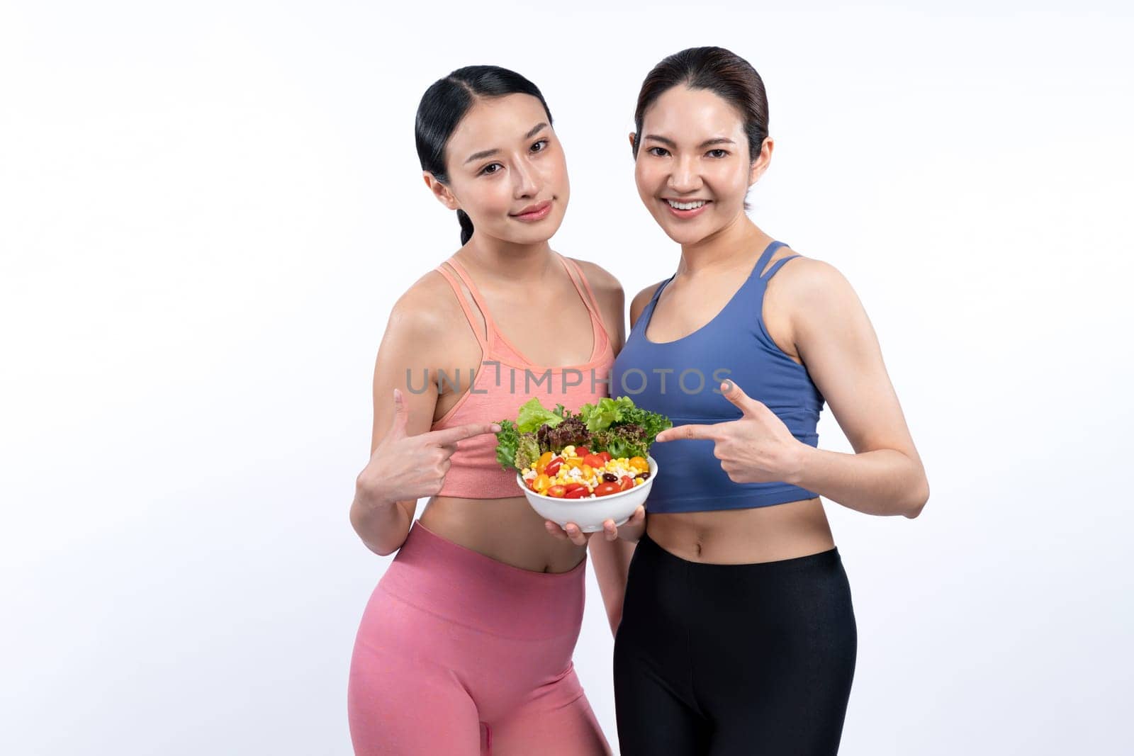 Two young sporty Asian women in sportswear holding salad bowl fill with fruit and vegetable. Natural youthful and fit body lifestyle people with balance nutrition on isolated background. Vigorous