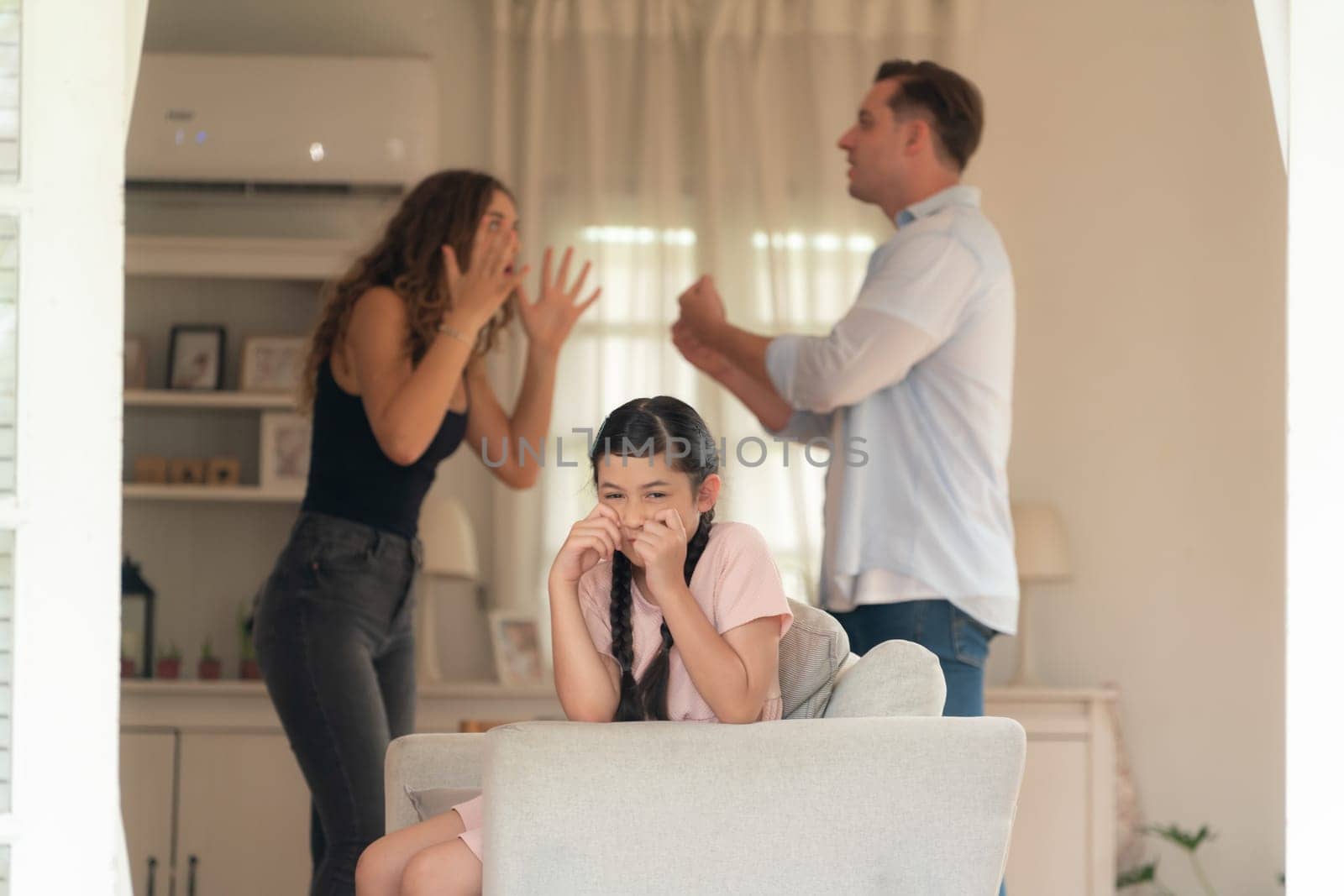 Annoyed and unhappy young girl sitting on sofa trapped in middle of tension by her parent argument in living room. Unhealthy domestic lifestyle and traumatic childhood develop to depression.Synchronos