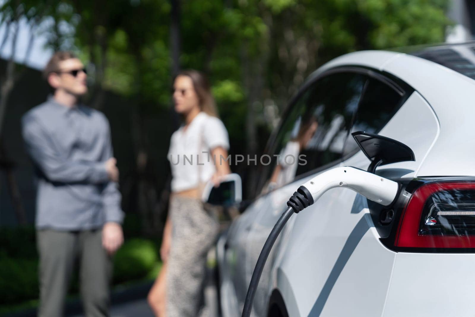 Young couple travel with EV electric car charging in green sustainable city outdoor garden in summer shows urban sustainability lifestyle by green clean rechargeable energy of electric vehicle innards