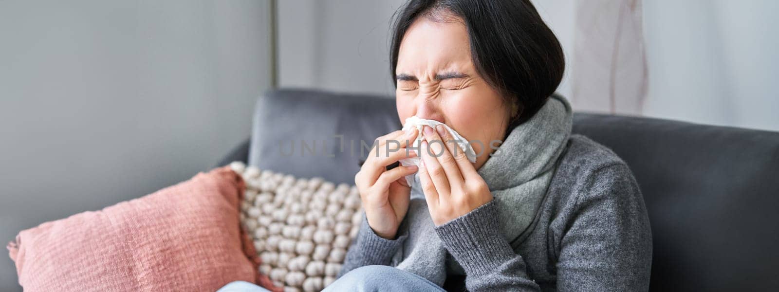 Close up of young korean woman staying at home with cold, sneezing in napking, has runny nose, concept of illness, health and influenza.