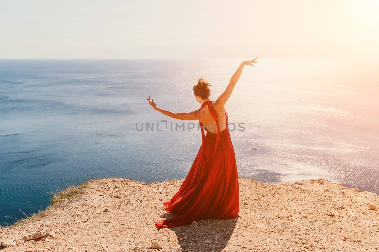 Side view a Young beautiful sensual woman in a red long dress posing on a rock high above the sea during sunrise. Girl on the nature on blue sky background. Fashion photo.