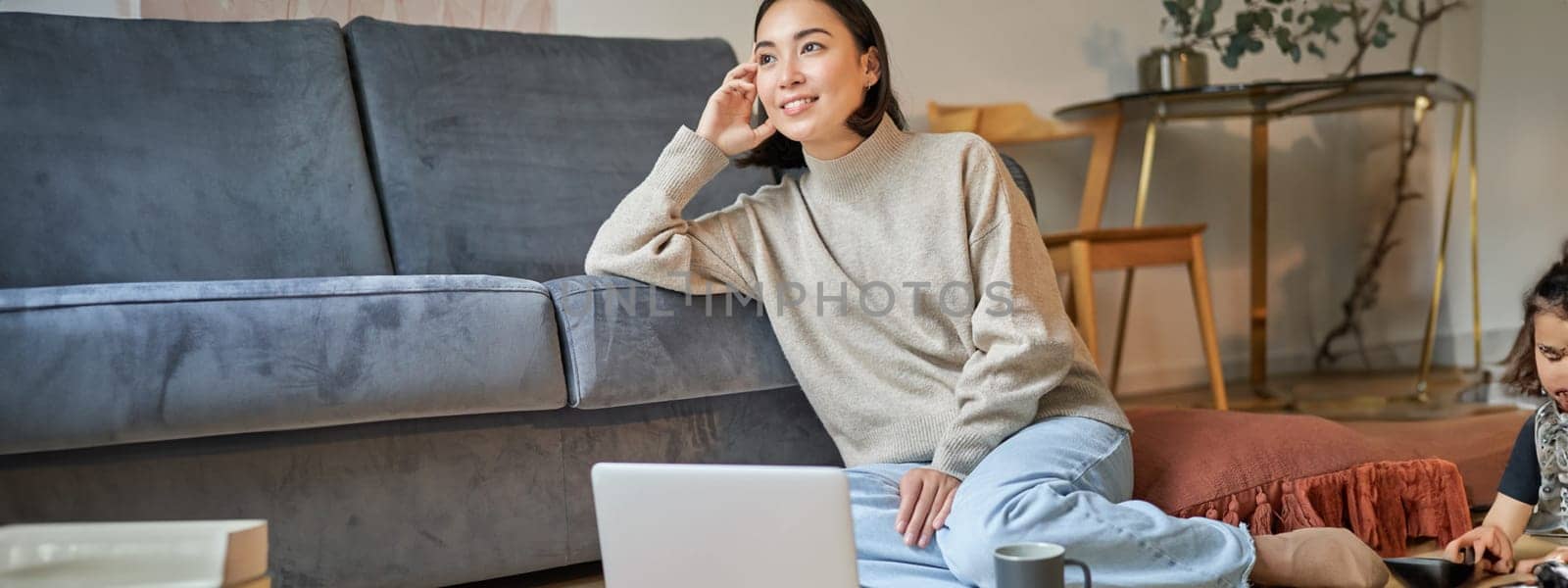 Image of smiling asian woman drinking hot tea, holding cup and sitting near laptop on floor, resting at home by Benzoix
