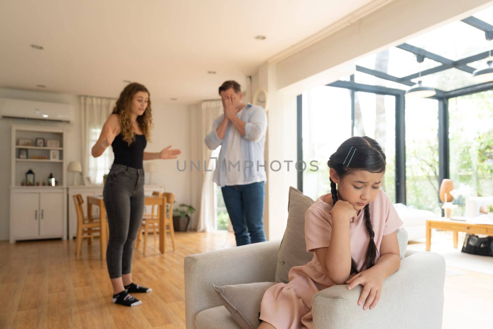 Annoyed and unhappy young girl sitting on sofa trapped in middle of tension by her parent argument in living room. Unhealthy domestic lifestyle and traumatic childhood develop to depression.Synchronos