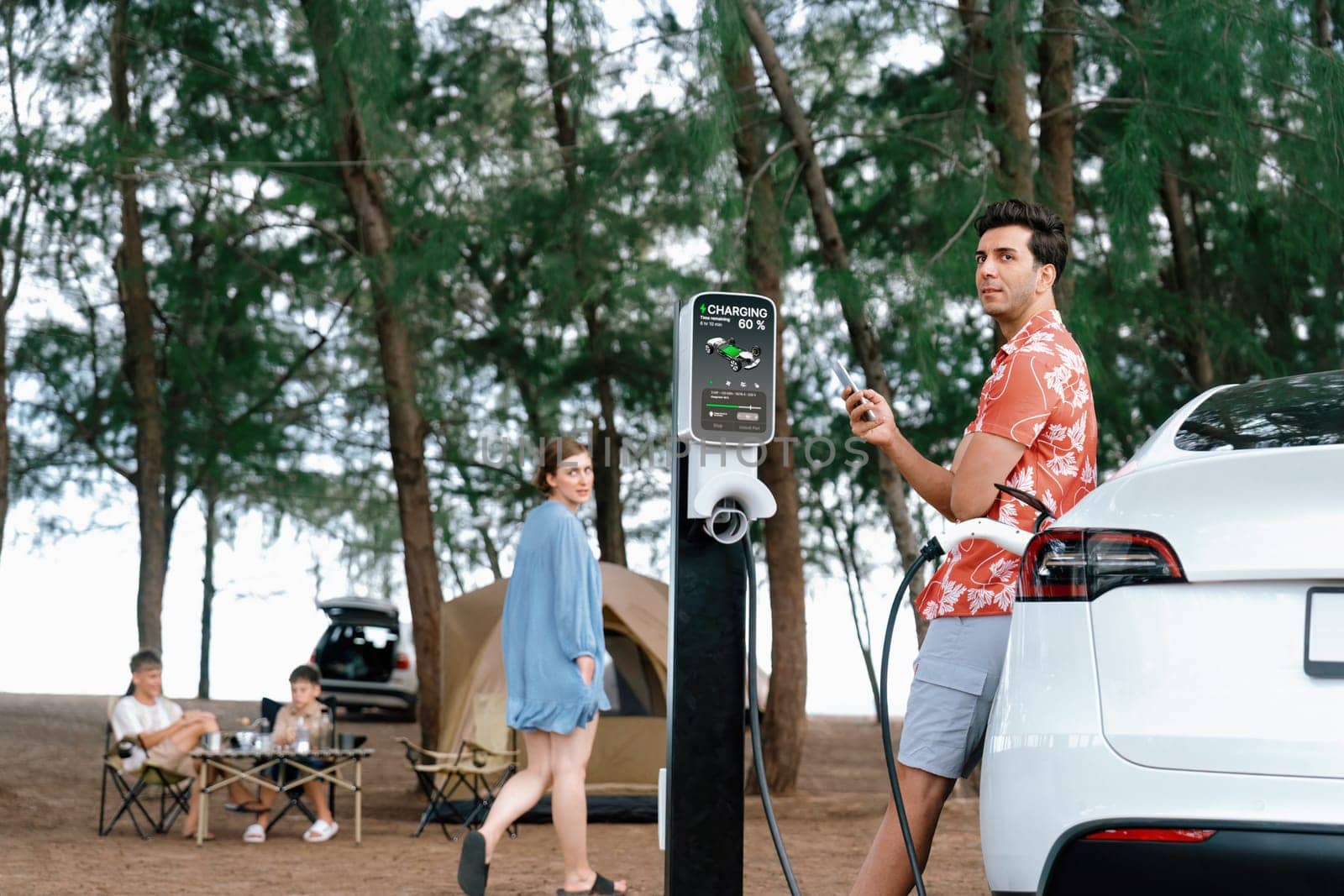 Outdoor adventure and family vacation camping in nature travel by eco friendly car for sustainable future. Lovely sit on trunk, charging EV car with EV charging station in campsite. Perpetual