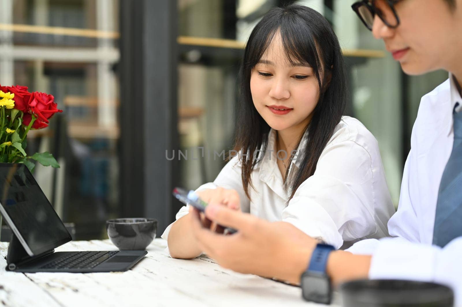Pretty young female office worker talking and watching social media content on mobile phone with colleague during coffee break.