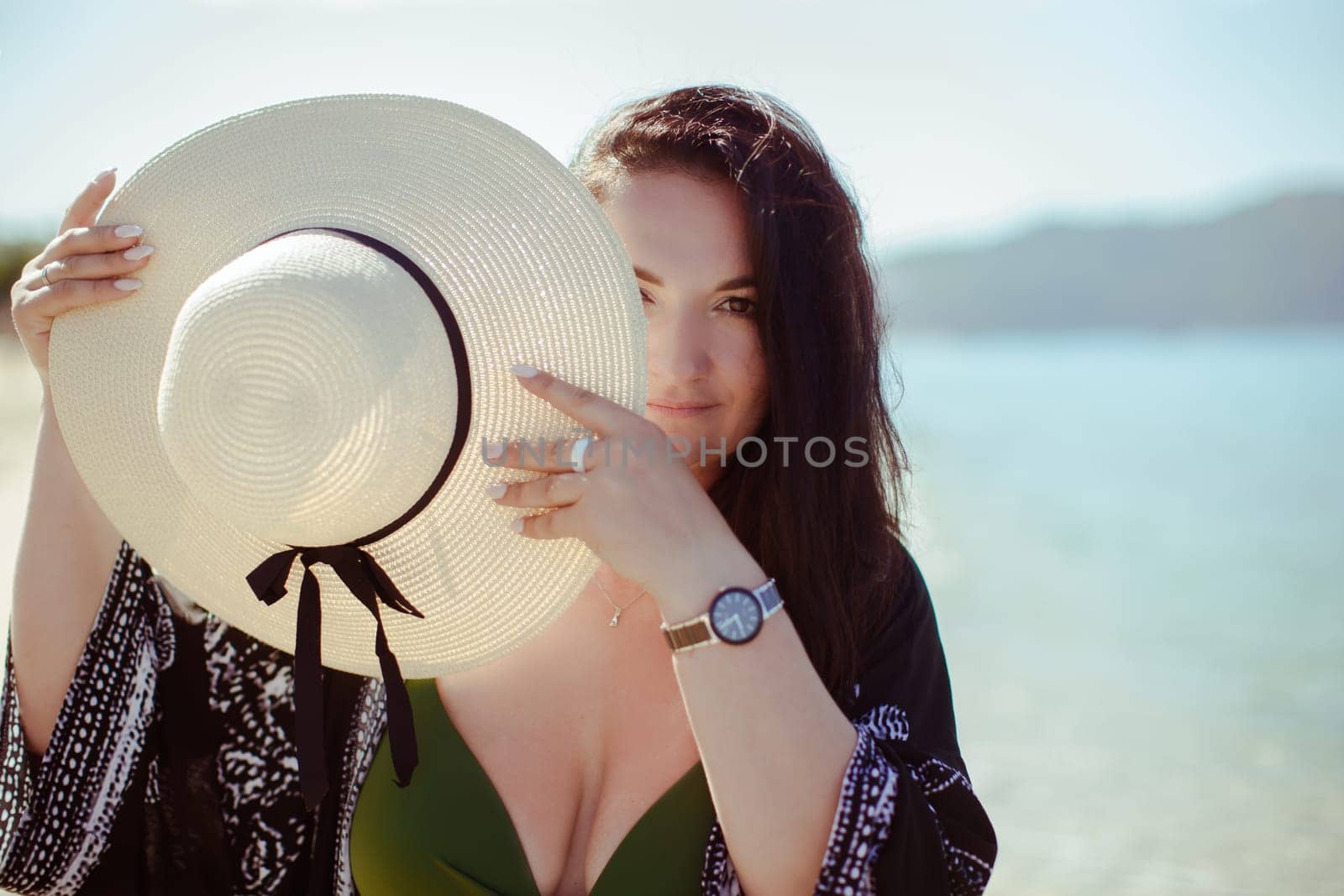 A brunette girl covers her face with a hat with a large brim in a black pareo against the background of the sea and mountains