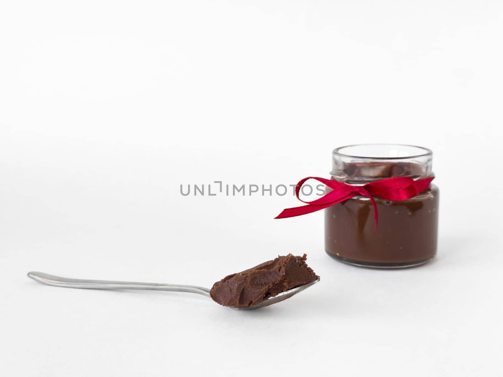 Elegant glass jar of hazelnut chocolate spread with red ribbon and spoonful on white background.
