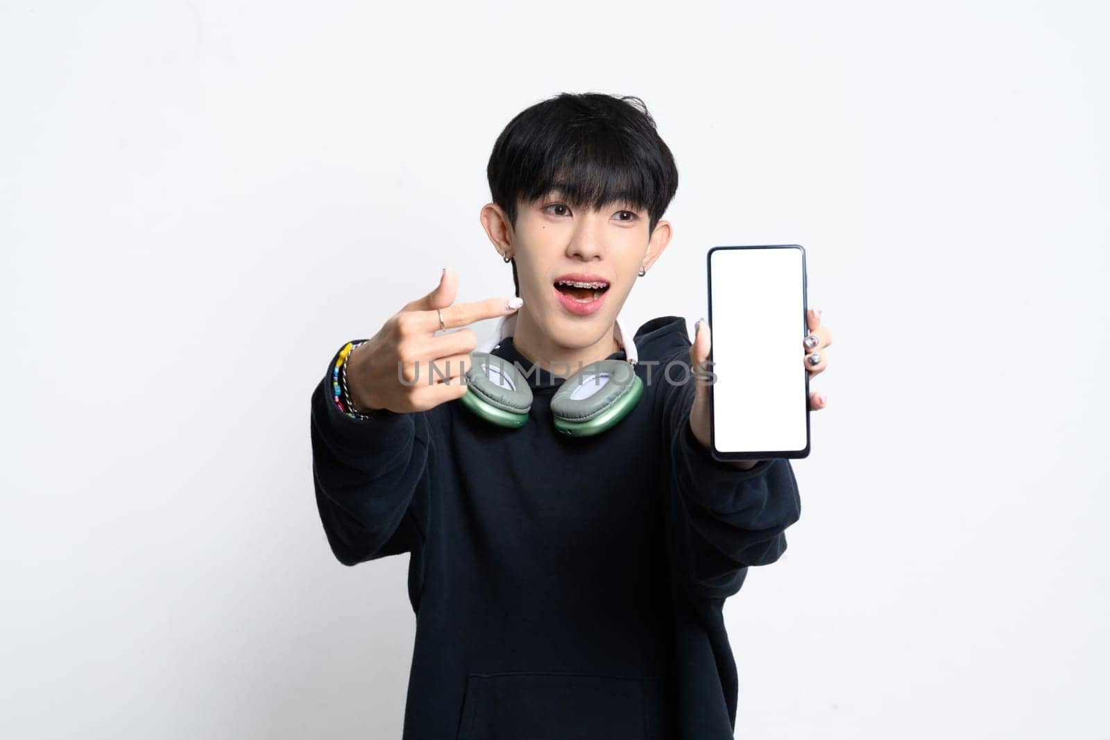 Cute young asian man in hoodie excitedly pointing at smartphone with a blank screen.
