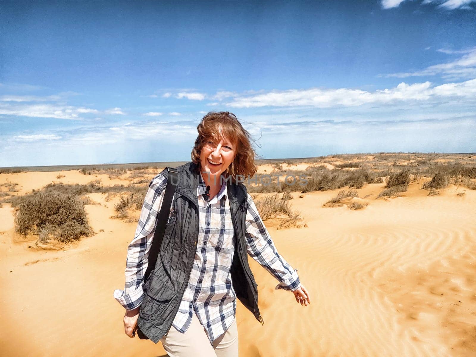 A cheerful middle-aged woman walks through a sunny desert, surrounded by golden sand dunes and a blue sky with clouds.