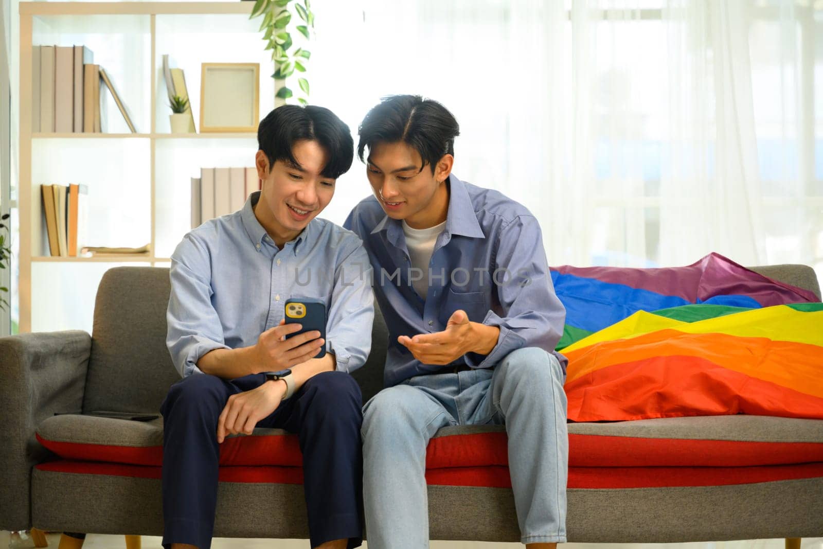 Young smiling gay couple using smartphone while sitting on a couch at home.