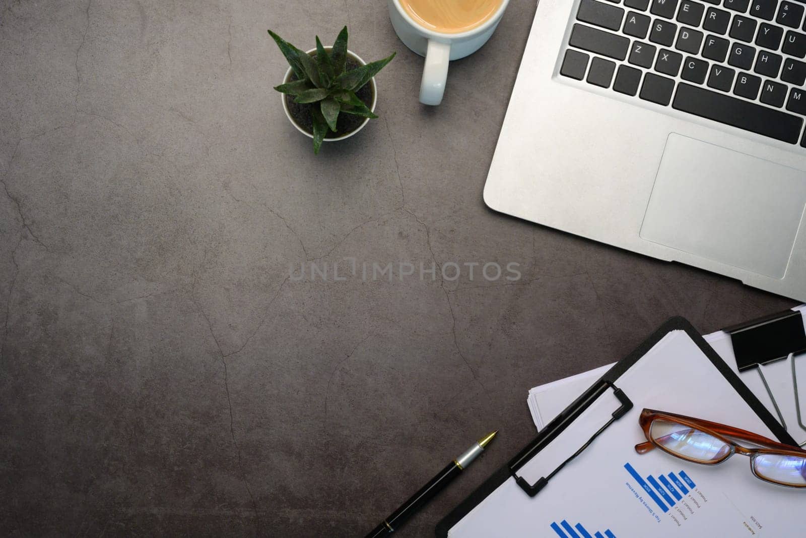 Top view of laptop, cup of coffee and financial reports on dark stone textured surface.