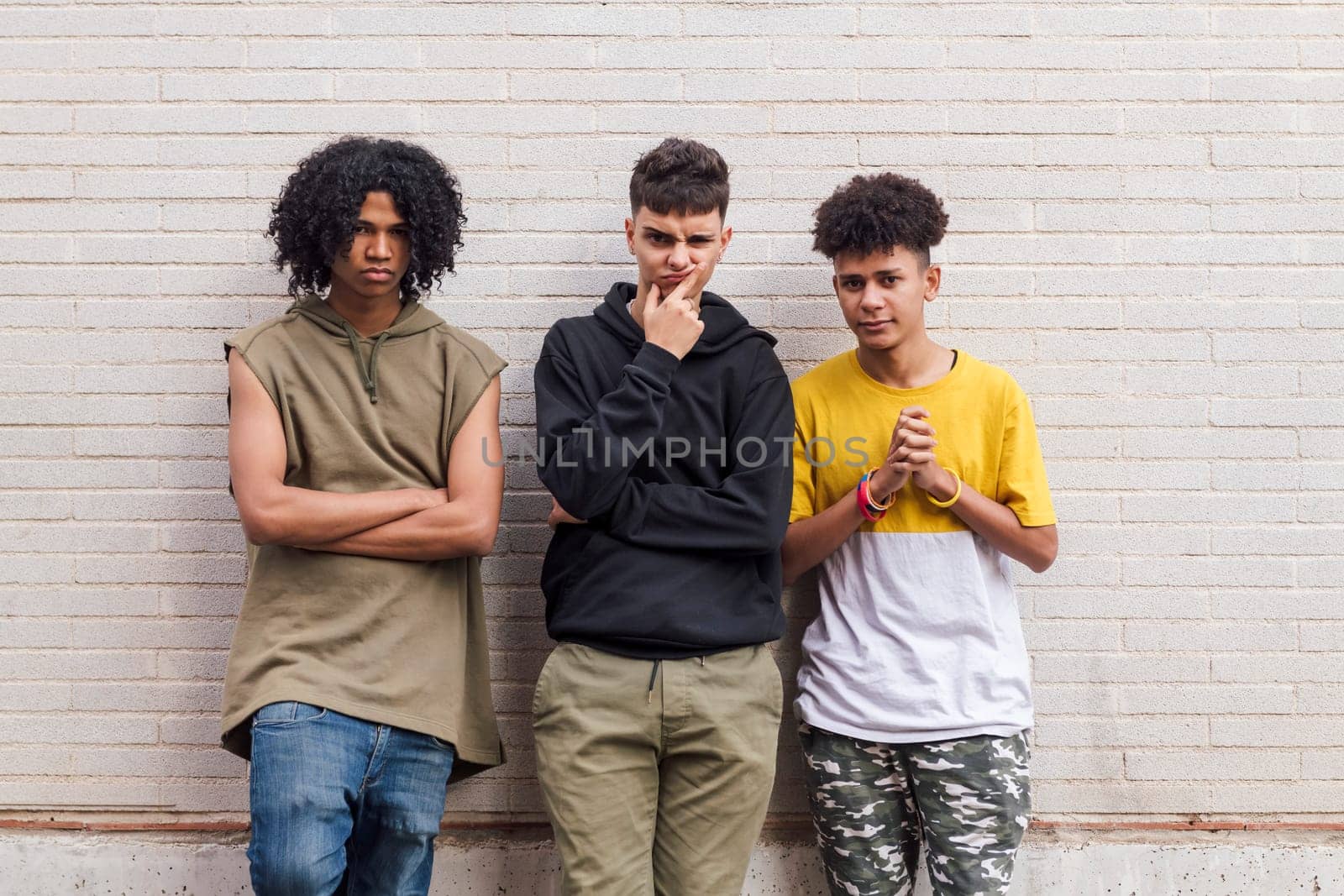 three multiethnic teenager friends with folded hands leaning against white brick wall, concept of friendship in youth and urban lifestyle