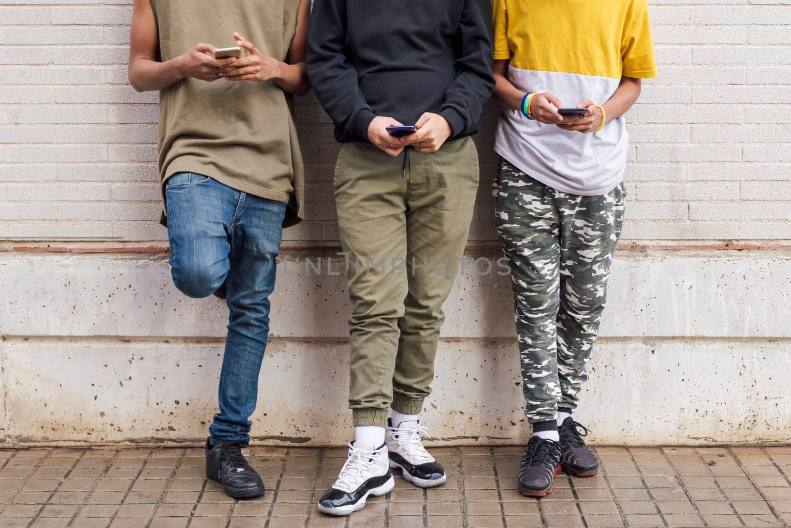crop of three unrecognizable teens addicted to mobile phones browsing Internet while leaning on brick wall, concept of youth and technology of communication