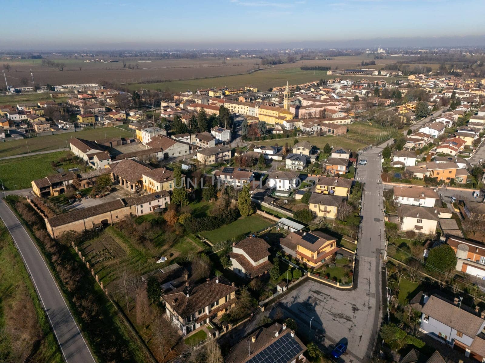 Aerial view capturing the small town of Stagno Lombardo in Cremona, Italy, featuring residential houses with solar panels, a church, and cultivated fields under a clear blue sky