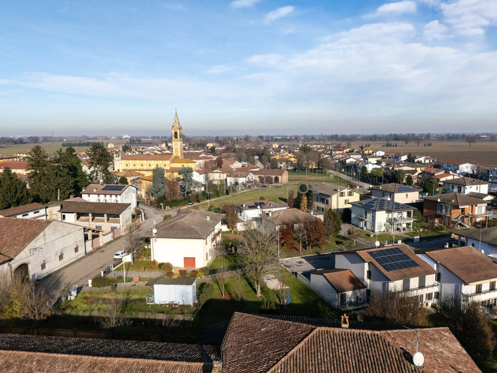 Aerial view capturing the small town of Stagno Lombardo in Cremona, Italy, featuring residential houses with solar panels, a church, and cultivated fields under a clear blue sky