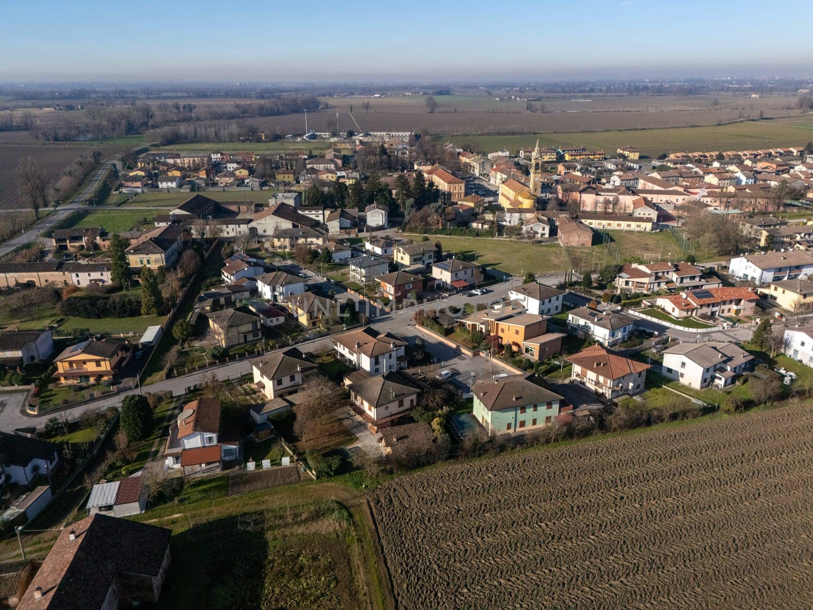 Aerial view of Stagno Lombardo, a small town in the province of Cremona, Lombardy, Italy, showcasing residential buildings, farmlands, and the surrounding countryside under a clear blue sky
