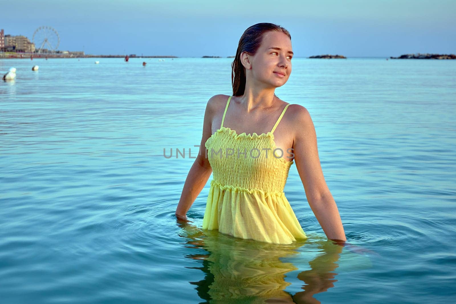 Carefree woman wearing yellow sundress enjoying freedom in the calm sea at sunset.