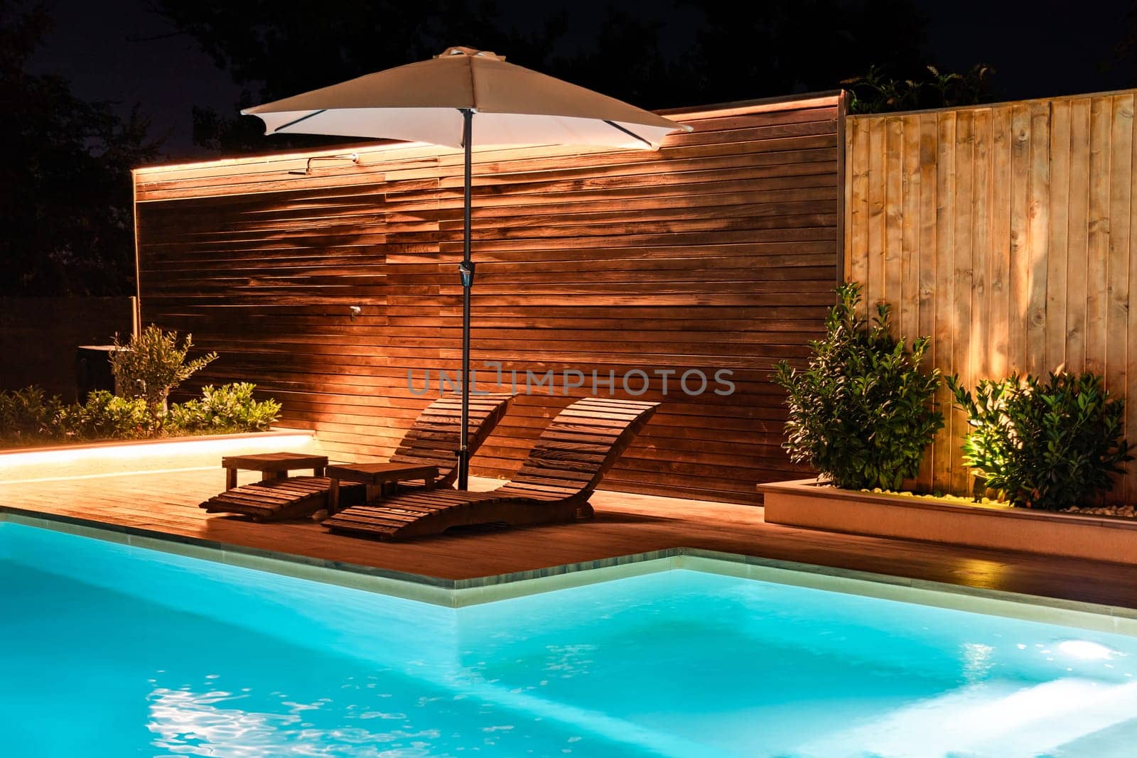 Elegant swimming pool with lounge chairs and umbrella at night. Luxury outdoor space illuminated with warm lights