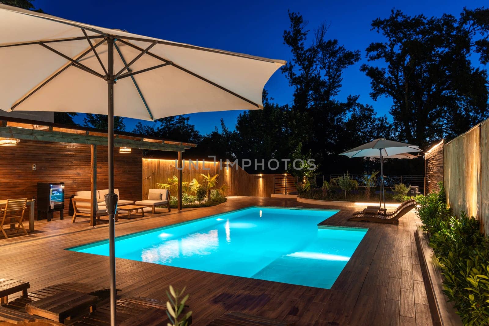 Backyard swimming pool with wooden deck and stylish umbrella under evening sky. Modern outdoor lounge area with serene ambiance and elegant lighting