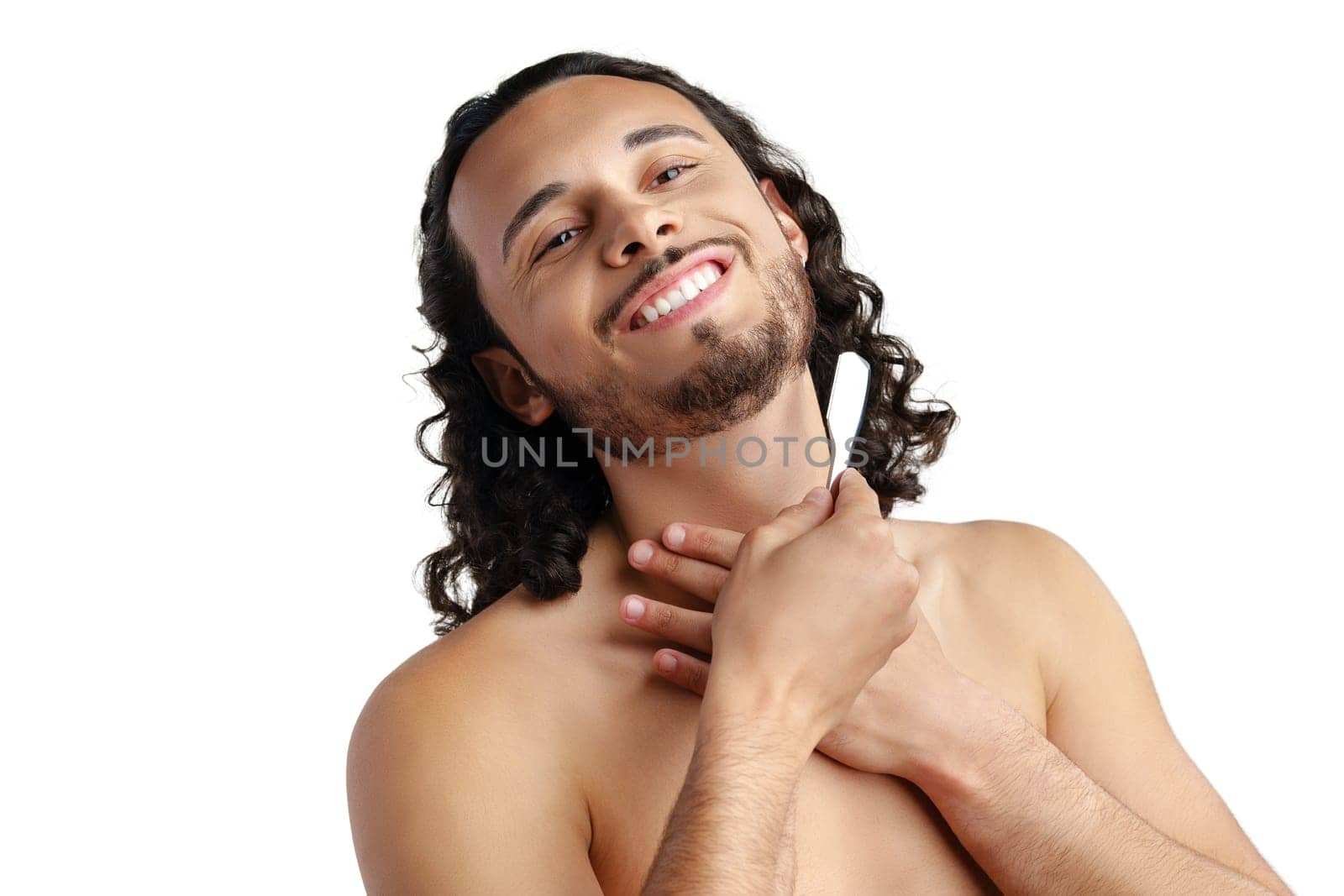 A man with curly hair grooming his beard in a well-lit space while using a straight razor for a clean shave