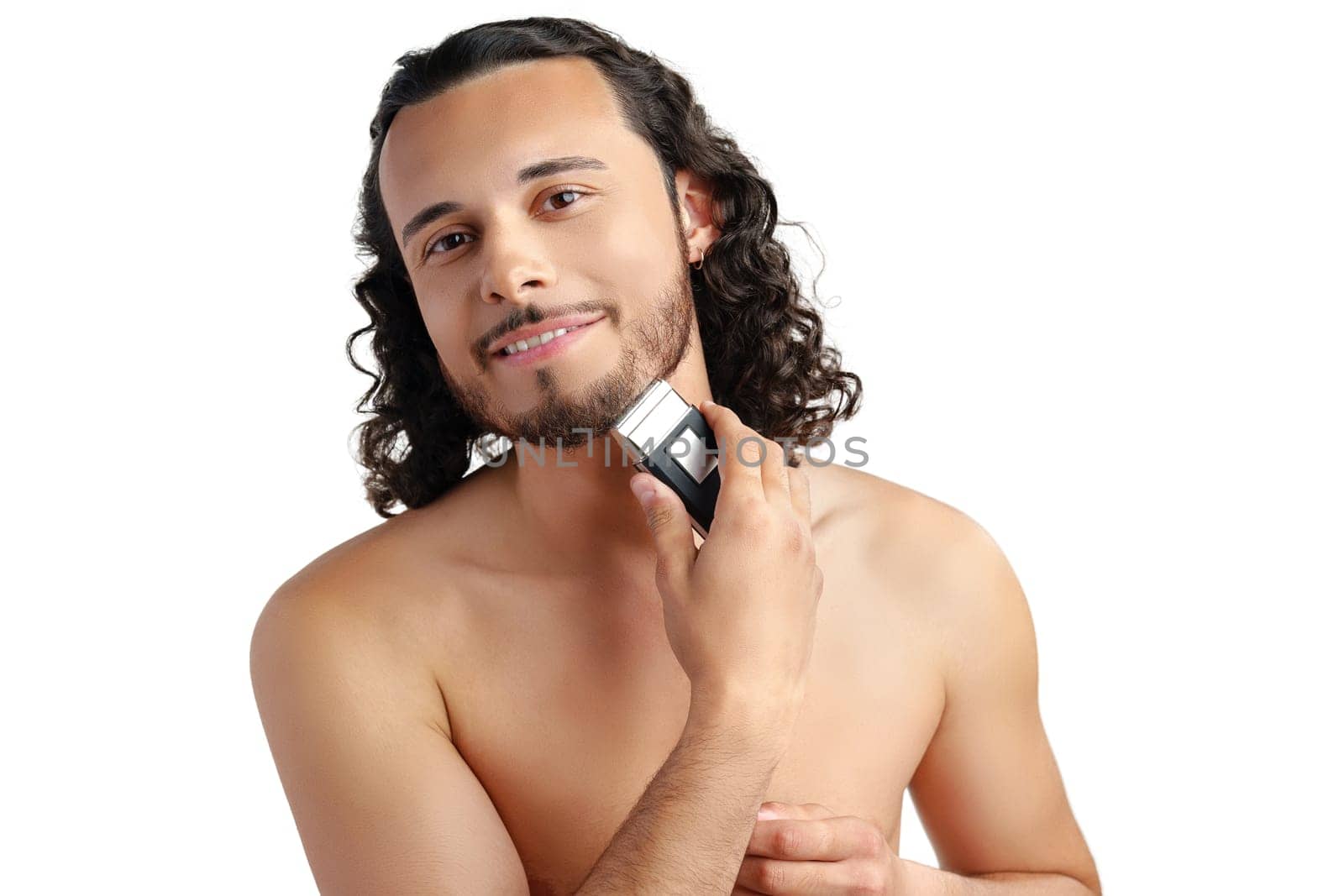 A young man with curly hair grooming his beard using an electric trimmer in a well-lit indoor space