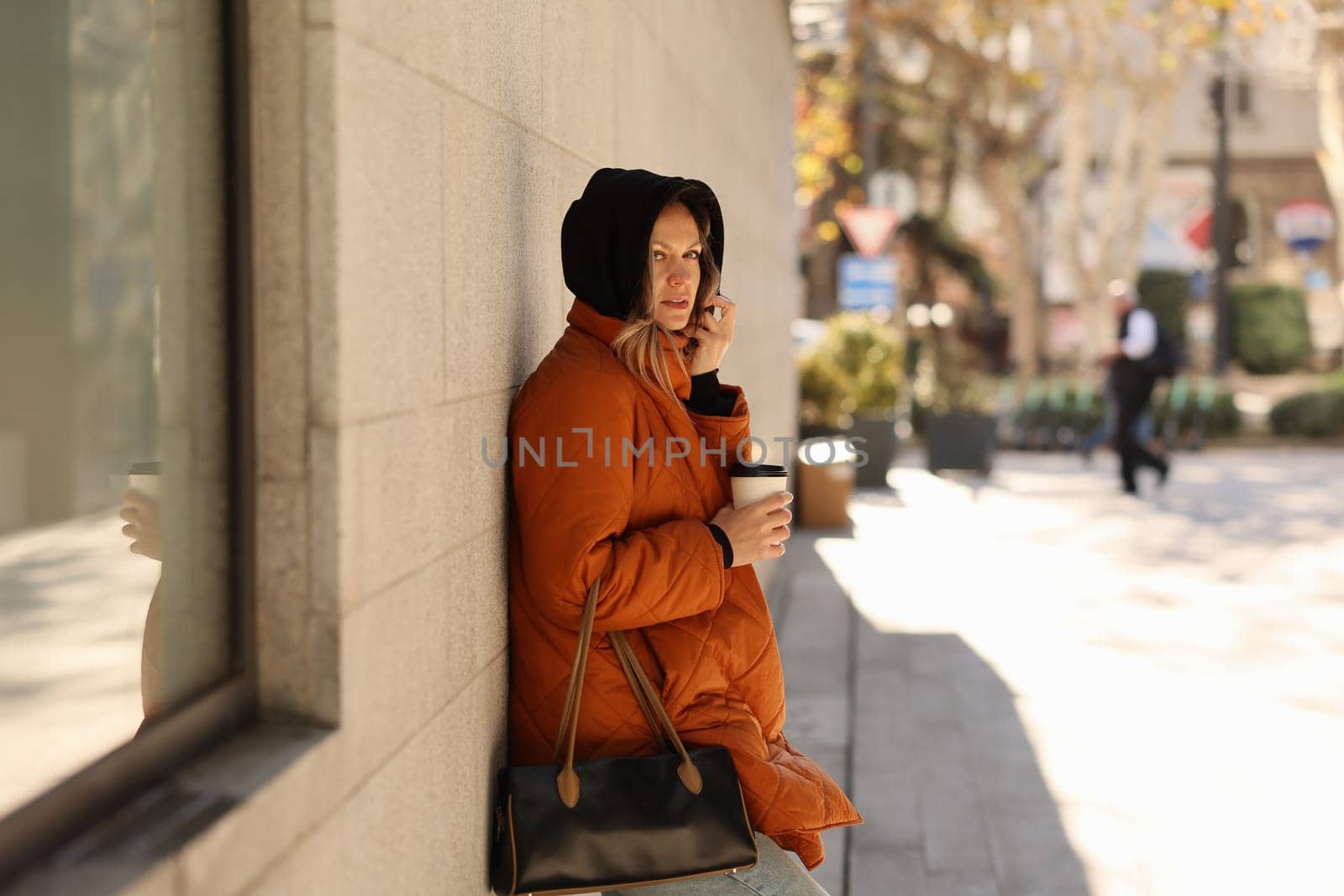 A woman in a bright orange coat holds a warm coffee beside a wall as the busy city vibrantly moves around her