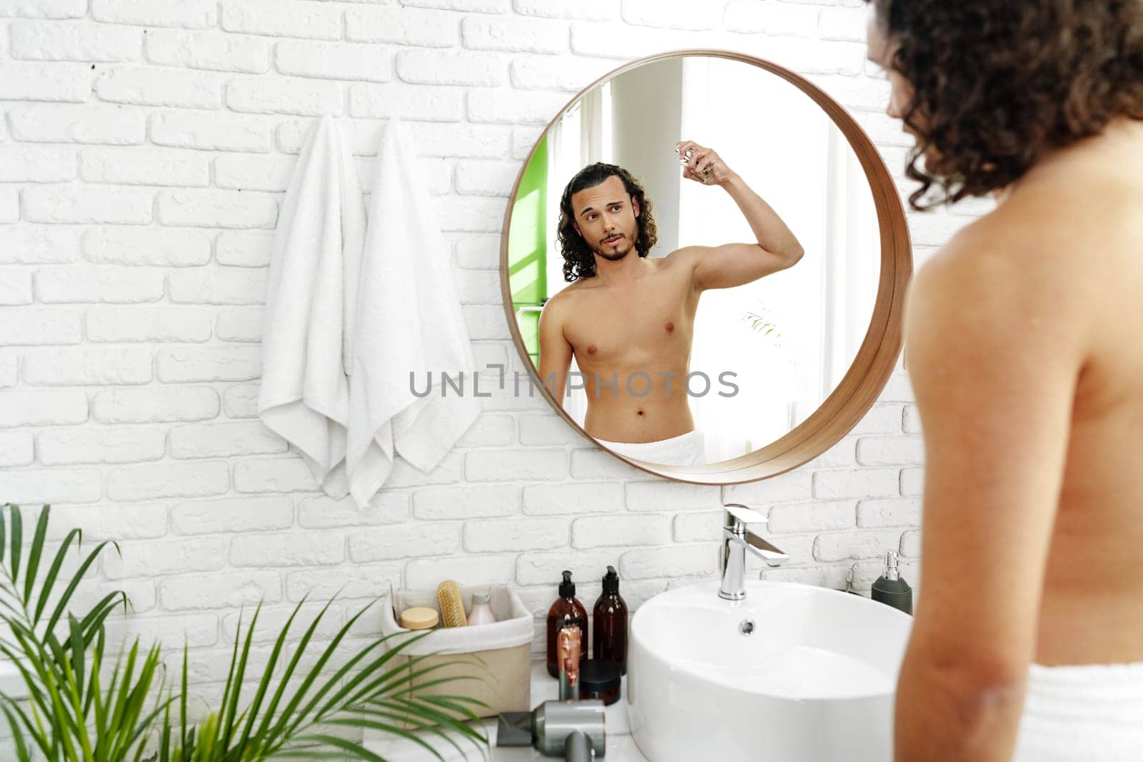 Man applying fragrance while looking in a round mirror in a bright bathroom close up
